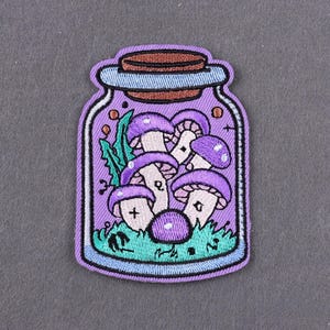 May include: Embroidered patch showing a jar filled with purple mushrooms. The jar has a light purple border and a brown cork lid. The mushrooms have white stems and purple caps with white spots. Green grass and leaves are at the bottom of the jar.