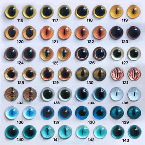 May include: A collection of glass eye cabochons in various colors and designs. The eyes range from yellow and gold to orange, brown, and blue, with black pupils. Each eye is numbered, with the numbers 116 through 143 visible.