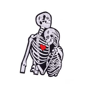 Op de afbeelding: Geborduurde patch met twee omarmende skeletten. De skeletten zijn wit met zwarte contouren en details, waaronder een rood hart. De patch is ontworpen voor toepassing op kleding of accessoires, met afmetingen in centimeters.