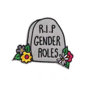 May include: A grey patch shaped like a tombstone, featuring the black text "R.I.P GENDER ROLES". The patch is adorned with colourful flowers in yellow, pink, and white, with green leaves. The patch is set against a white backdrop.