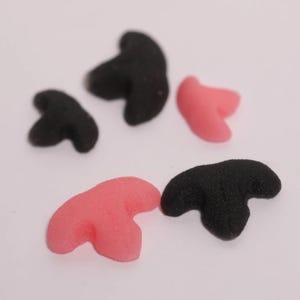 May include: Close-up of several black and pink foam clown noses. The noses are in various sizes and orientations, with a textured surface. The background is a plain white surface.
