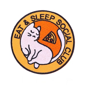 May include: Embroidered patch featuring a white cat curled up within a circle. The cat is set against an orange background with a pizza slice. The patch has the words "EAT & SLEEP SOCIAL CLUB" in a yellow border.