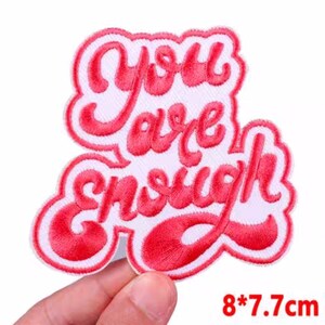 May include: A red embroidered patch with the words "You are enough" in a cursive font. The patch has a white border and measures 8 inches by 7.7 inches.