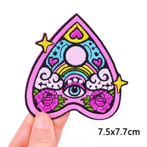 May include: A pink, heart-shaped embroidered patch with a decorative design. The patch features a rainbow, eye, hearts, stars, and roses. The patch measures 7.5 x 7.7 cm.