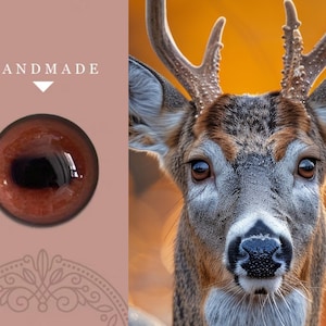 May include: Close-up of a handmade brown glass eye with a black pupil. The eye is set against a pink background with the word "HANDMADE" in white text above it.