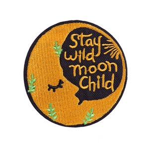 Op de afbeelding: Geborduurde ronde patch met een zwart en goud ontwerp. De patch heeft de tekst "Stay Wild Moon Child" in een gestileerd lettertype, tegen een halve maan vorm. Een kleine zwarte hond en groene bladeren zijn ook in het ontwerp opgenomen.