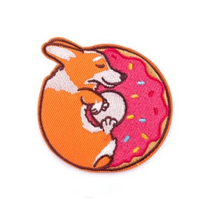 Puede incluir: Parche bordado con un perro corgi de dibujos animados abrazando una rosquilla rosa con chispas de colores. El corgi es naranja y blanco, y la rosquilla tiene un contorno marrón. El parche es circular y tiene un borde naranja.