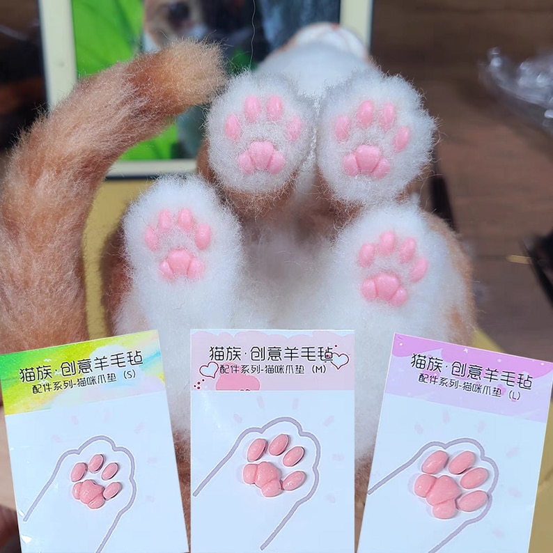 May include: A close-up of a cat's paws with pink paw pads and white fur. Three cards with pink paw-shaped pads are in front of the cat's paws. The cards have Chinese text and the sizes S, M, and L.