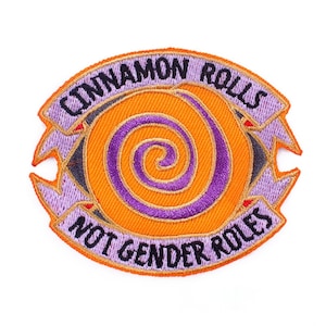 Puede incluir: Parche bordado con un diseño de espiral naranja y morado, rodeado por las palabras "CINNAMON ROLLS" y "NOT GENDER ROLES". El parche tiene un borde morado con detalles en naranja.