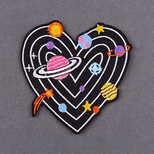 Puede incluir: Parche bordado en forma de corazón negro con contorno blanco, que representa un sistema solar colorido. El parche incluye planetas, estrellas y un cometa, ideal para personalizar ropa o accesorios.