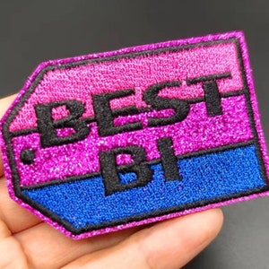 May include: A pink, blue, and black embroidered patch with the text "BEST BI" in black lettering. The patch has a glittery finish.