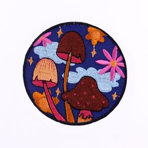 May include: Embroidered patch featuring a whimsical design of three mushrooms in shades of brown and orange, set against a dark blue background with light blue clouds, pink flowers, and gold stars. The patch is round.