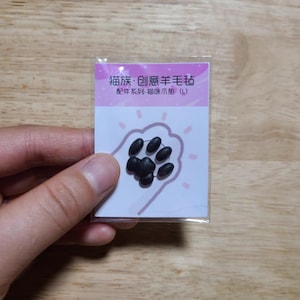 May include: A small, clear plastic package containing black cat paw-shaped felt pieces. The package has text in Chinese characters and a pink background with a paw print illustration. The felt pieces are arranged to resemble a cat's paw.