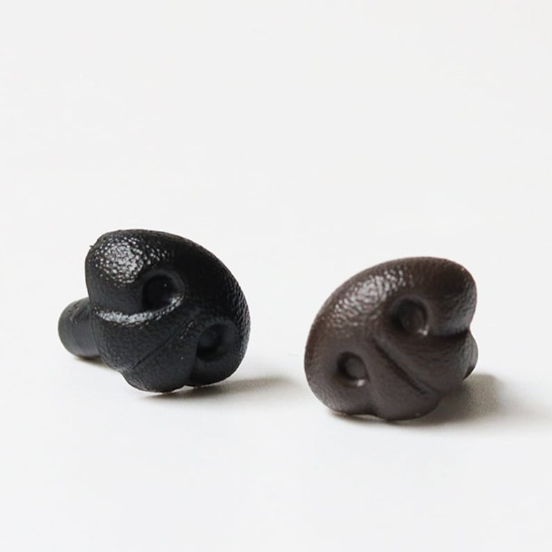 May include: Two plastic dog noses, one black and one brown, are displayed against a white background. The noses have a textured surface and are designed with nostril details. These are likely craft supplies or costume accessories.
