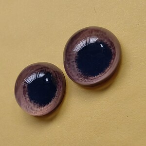 K&ouml;nnte beinhalten: Zwei Glas-Puppenaugen mit einer dunkelbraunen Iris und einem helleren braunen Au&szlig;enring. Die Augen haben eine gl&auml;nzende Oberfl&auml;che und reflektieren das Licht. Der Hintergrund ist einfarbig gelb.
