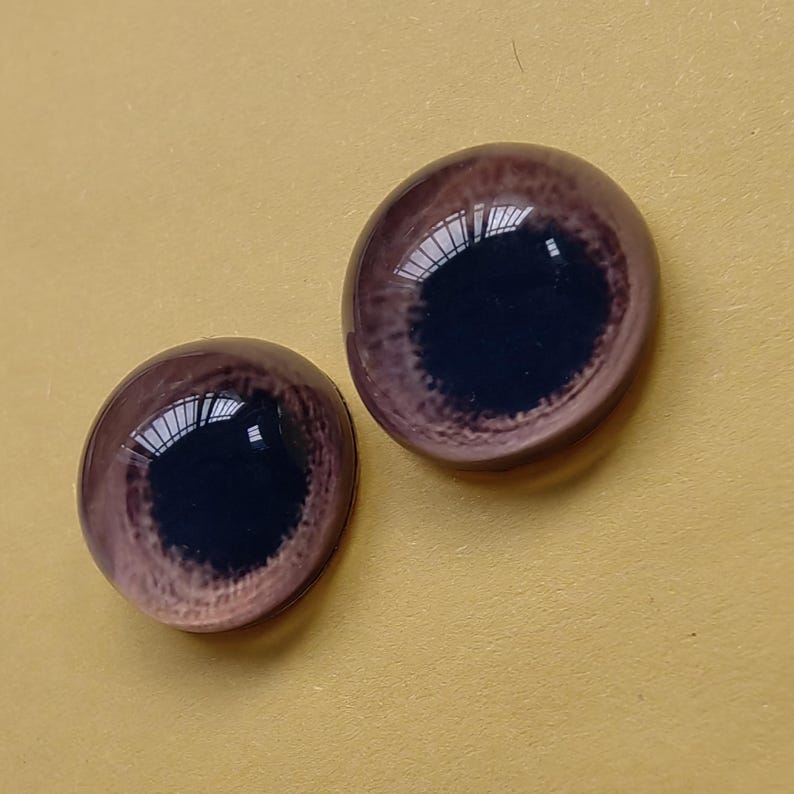 K&ouml;nnte beinhalten: Zwei Glas-Puppenaugen mit einer dunkelbraunen Iris und einem helleren braunen Au&szlig;enring. Die Augen haben eine gl&auml;nzende Oberfl&auml;che und reflektieren das Licht. Der Hintergrund ist einfarbig gelb.