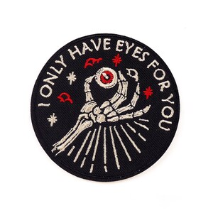 Può includere: Una toppa rotonda ricamata nera con la scritta "I ONLY HAVE EYES FOR YOU". Presenta una mano scheletrica che tiene un occhio, con dettagli rossi e bianchi, circondata da stelle e altri piccoli accenti rossi.