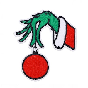 以下が含まれることがあります： 緑色の手と白いトリム、赤い袖が赤いクリスマスオーナメントを持っている刺繍パッチ。パッチは白い縁取りがあり、ホリデーをテーマにした工芸品やアパレル向けに設計されています。