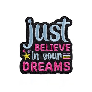 以下が含まれることがあります： 「just believe in your dreams」の文字が水色とピンクで刺繍されたパッチ。パッチは黒い縁取りで、黄色の星と紫の花が特徴です。