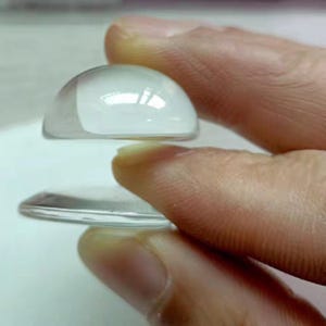 May include: Two clear glass cabochons, one domed and one flat, are held between fingers. The domed cabochon reflects light, while the flat one is a clear disc. The image highlights the transparency and shape of the glass, suitable for crafts.