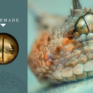 K&ouml;nnte beinhalten: Nahaufnahme eines handgefertigten Glasauges in goldbrauner Farbe mit einer schwarzen vertikalen Pupille. Das Auge ist vor einem blauen Hintergrund mit dem Wort "HANDMADE" dar&uuml;ber platziert.