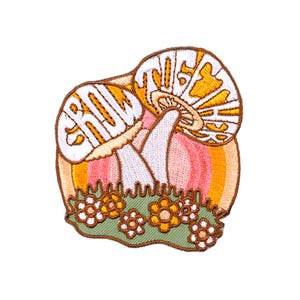 May include: Embroidered patch featuring two stylized mushrooms with the word "GROW" in white lettering. The mushrooms are set against a rainbow background with green grass and small white and yellow flowers.