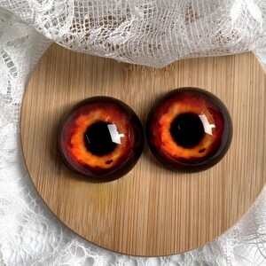 May include: Two large, round, red and orange glass eyes with black pupils. The eyes are on a wooden surface.