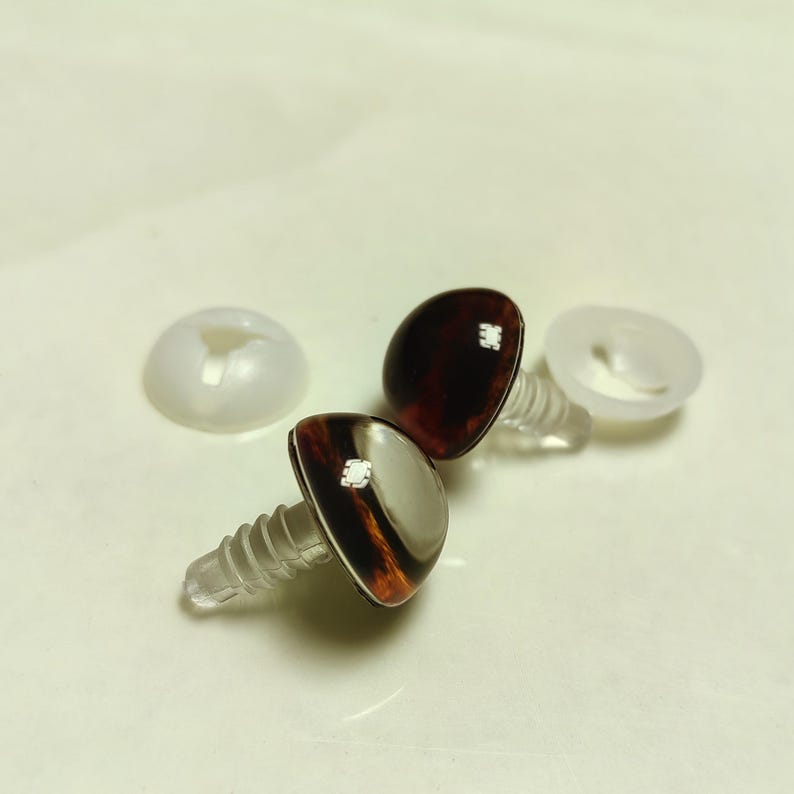 K&ouml;nnte beinhalten: Zwei braune und bernsteinfarbene Puppenaugen mit transparenten Kunststoffschraubpfosten und wei&szlig;en Kunststoff-R&uuml;ckenteilen. Die Augen haben eine gl&auml;nzende Oberfl&auml;che und ein detailliertes, realistisches Aussehen. Ideal f&uuml;r Basteln und Puppenherstellung.