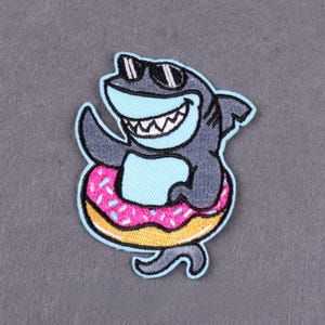 Puede incluir: Parche bordado con un tiburón de dibujos animados con gafas de sol y una gran sonrisa, flotando en una rosquilla rosa con chispas. El tiburón es gris y azul, con una boca azul claro y dientes blancos. La rosquilla es amarilla y rosa.