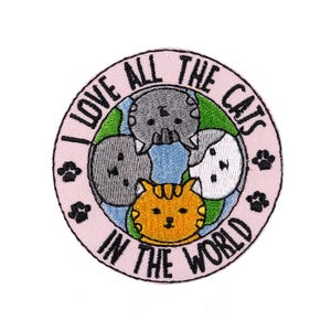 Può includere: Toppa ricamata con la scritta "I LOVE ALL THE CATS IN THE WORLD" in un cerchio rosa. La toppa presenta quattro gatti dei cartoni animati in grigio, bianco e arancione, con accenti verdi e blu. Impronte di zampe nere all'esterno del cerchio.