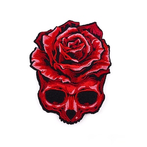 May include: Embroidered patch featuring a red skull with a red rose on top. The skull has black eye sockets and a black nose. The rose is detailed with multiple layers of red petals. The patch is likely for clothing or accessories.
