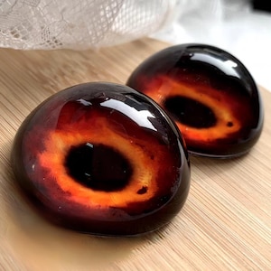 May include: Two round, black resin cabochons with a red and orange ring around a black centre. The cabochons are glossy and have a 3D effect.