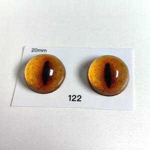 May include: Two glass cabochons with a cat eye design. The eyes are amber with a black vertical slit pupil. The cabochons are 20mm in diameter and are on a white card with the number 122.