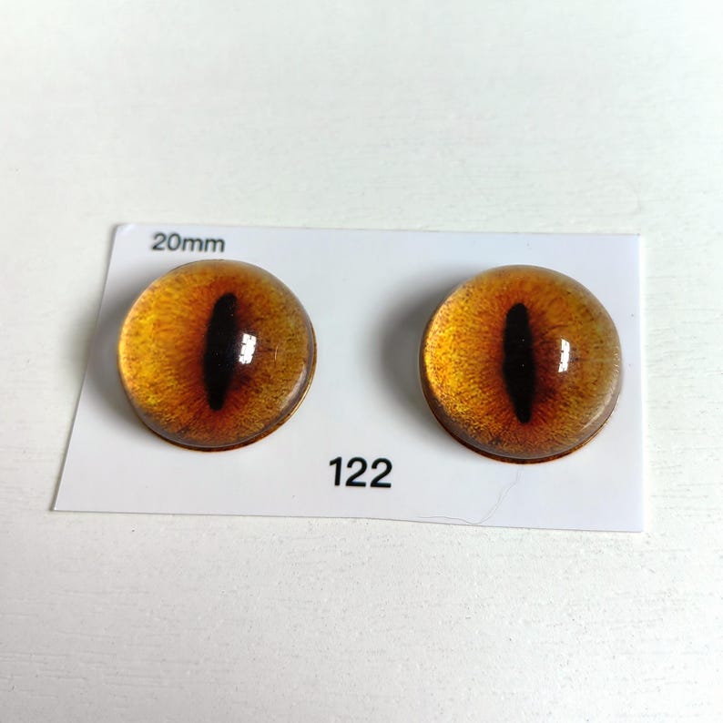 May include: Two glass cabochons with a cat eye design. The eyes are amber with a black vertical slit pupil. The cabochons are 20mm in diameter and are on a white card with the number 122.