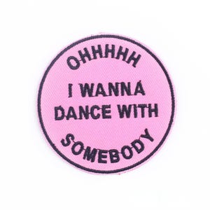 May include: A round, pink embroidered patch with the phrase "OHHHHH I WANNA DANCE WITH SOMEBODY" in black lettering. The patch has a black border and is set against a white background.