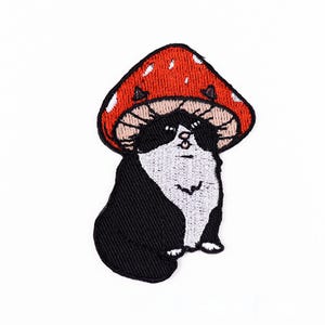 May include: Embroidered patch featuring a black and white cat wearing a red mushroom cap with white spots. The patch has a whimsical, cartoonish design, perfect for adding to clothing or accessories.