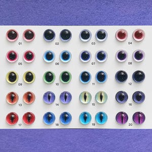 Puede incluir: Una colección de veinte pares de ojos de muñeca de cristal en varios colores, dispuestos en filas. Los colores incluyen rosa, azul, verde, rojo y morado. Cada ojo tiene una pupila negra. Números 01-20 impresos.
