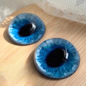 May include: Two resin eye cabochons with a realistic blue iris and a dark pupil. The irises have intricate details with shades of blue and white, creating a lifelike appearance. The cabochons are set against a light wooden surface.