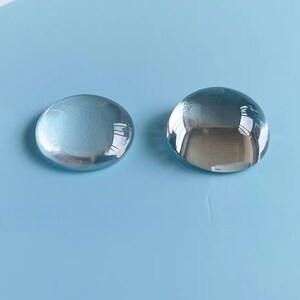May include: Two clear, round glass cabochons with a subtle blue hue, reflecting the surroundings. They sit on a light blue surface, creating a minimalist look. These could be used for jewellery making or other craft projects.