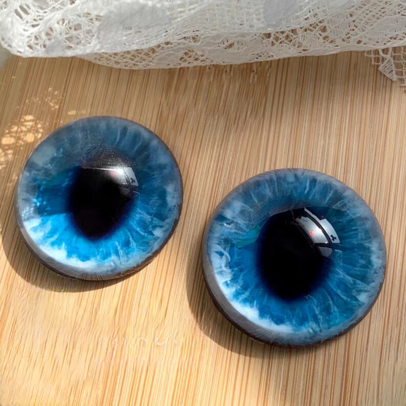 May include: Two round, glass eye cabochons with a deep blue iris and a black pupil. The iris has a detailed, swirling pattern with lighter blue and white accents. The eyes are set against a wooden surface.