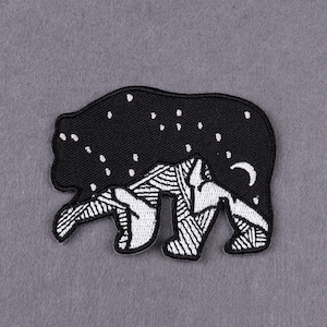 May include: Black and white embroidered bear patch. The bear's body is black with white stars and a crescent moon. The bear's legs and lower body are white with a geometric pattern.