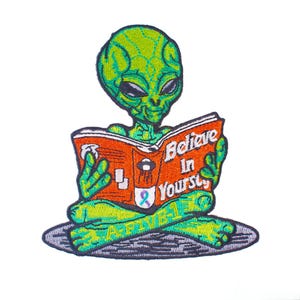 Op de afbeelding: Geborduurde patch met een groene alien die een boek leest met de tekst "Believe In Yourself" op de kaft. De alien zit met gekruiste benen. De patch heeft een grijze rand en de tekst "A-FIVE-1".