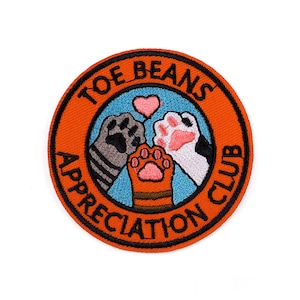 May include: Circular orange embroidered patch with the text "TOE BEANS APPRECIATION CLUB" in black. The patch features three cat paws in grey, black and white, and orange, with a pink heart above them. A fun accessory.