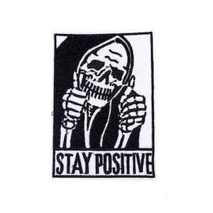 Pode incluir: Patch bordado a preto e branco com um esqueleto com capuz, fazendo um gesto de polegar para cima. A frase "STAY POSITIVE" está impressa em branco na parte inferior. O patch é retangular com uma borda preta, para roupas ou acessórios.