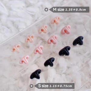Può includere: Una collezione di ciondoli a forma di naso in miniatura, rosa e neri. La taglia M misura 1,35 x 0,9 cm, e la taglia S misura 1,15 x 0,75 cm. Questi piccoli ciondoli dettagliati sono disposti su una superficie trasparente.