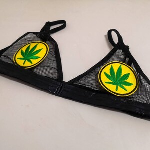 Puede incluir: Bralette de malla negra con un parche de hoja de cannabis en cada copa. Los parches presentan una hoja de cannabis verde sobre un fondo amarillo. El bralette tiene tirantes ajustables y una banda negra.