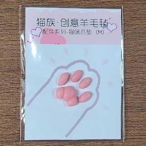 May include: A small, pink packet featuring a cat paw print design. Inside, there are several pink, oval-shaped items arranged to resemble a cat's paw. The packet has Chinese text and a heart graphic.