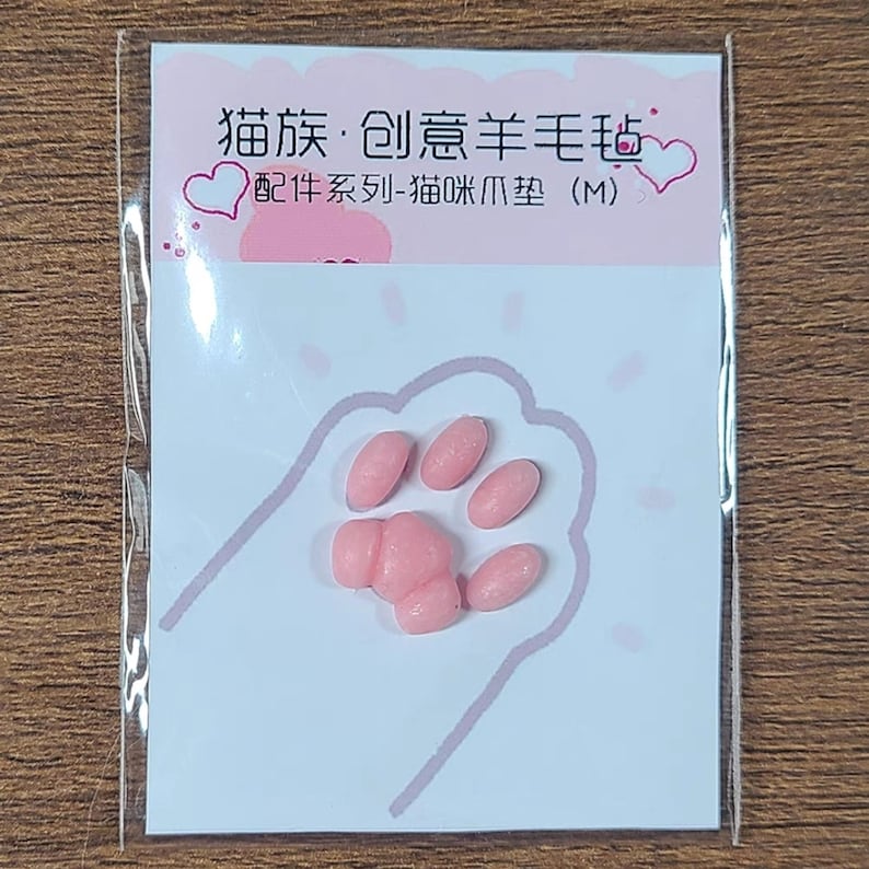 May include: A small, pink packet featuring a cat paw print design. Inside, there are several pink, oval-shaped items arranged to resemble a cat's paw. The packet has Chinese text and a heart graphic.