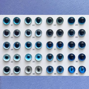 May include: A collection of thirty-two glass doll eyes in various shades of blue, arranged in rows. Each eye has a dark pupil and a glossy finish. The eyes are numbered from 21 to 40.