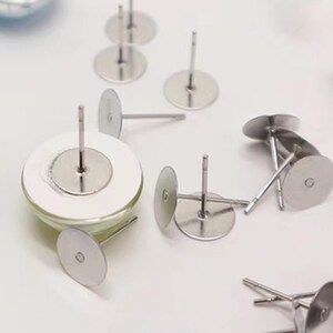 May include: Several silver earring posts with circular backings are scattered on a white surface. The posts are evenly spaced and appear to be made of metal. These components are likely for crafting or making jewellery, with a focus on earrings.